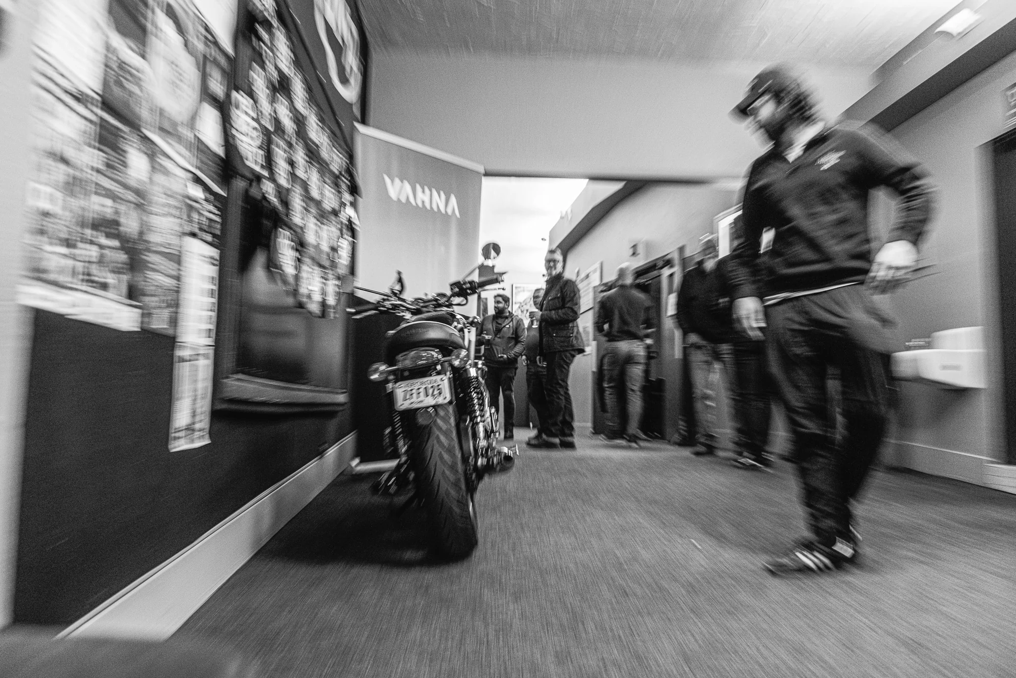 Black-and-white motion photograph of a motorcycle moving through the cinema lobby as people watch.
