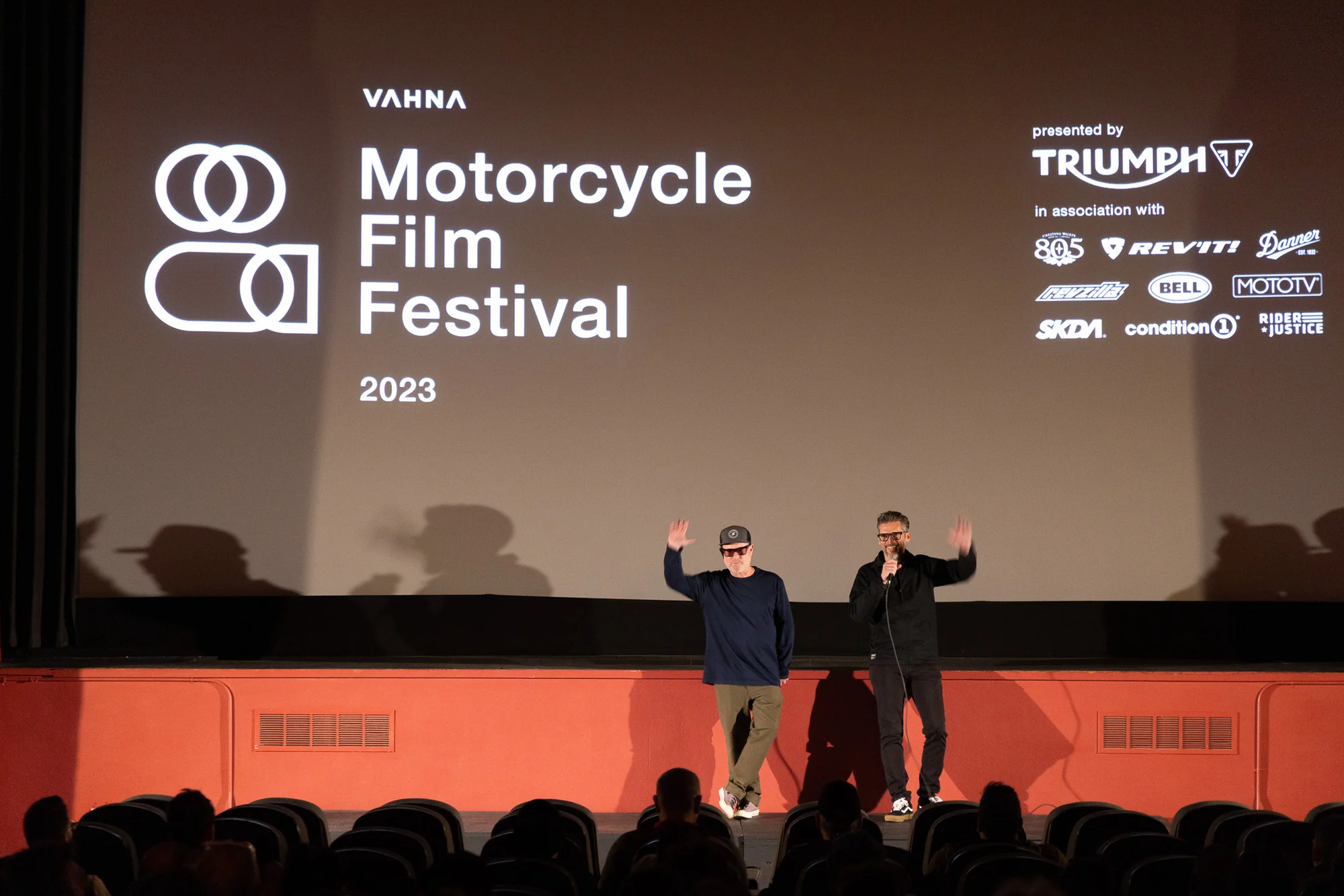Two presenters addressing the audience beneath the VAHNA Motorcycle Film Festival screen.