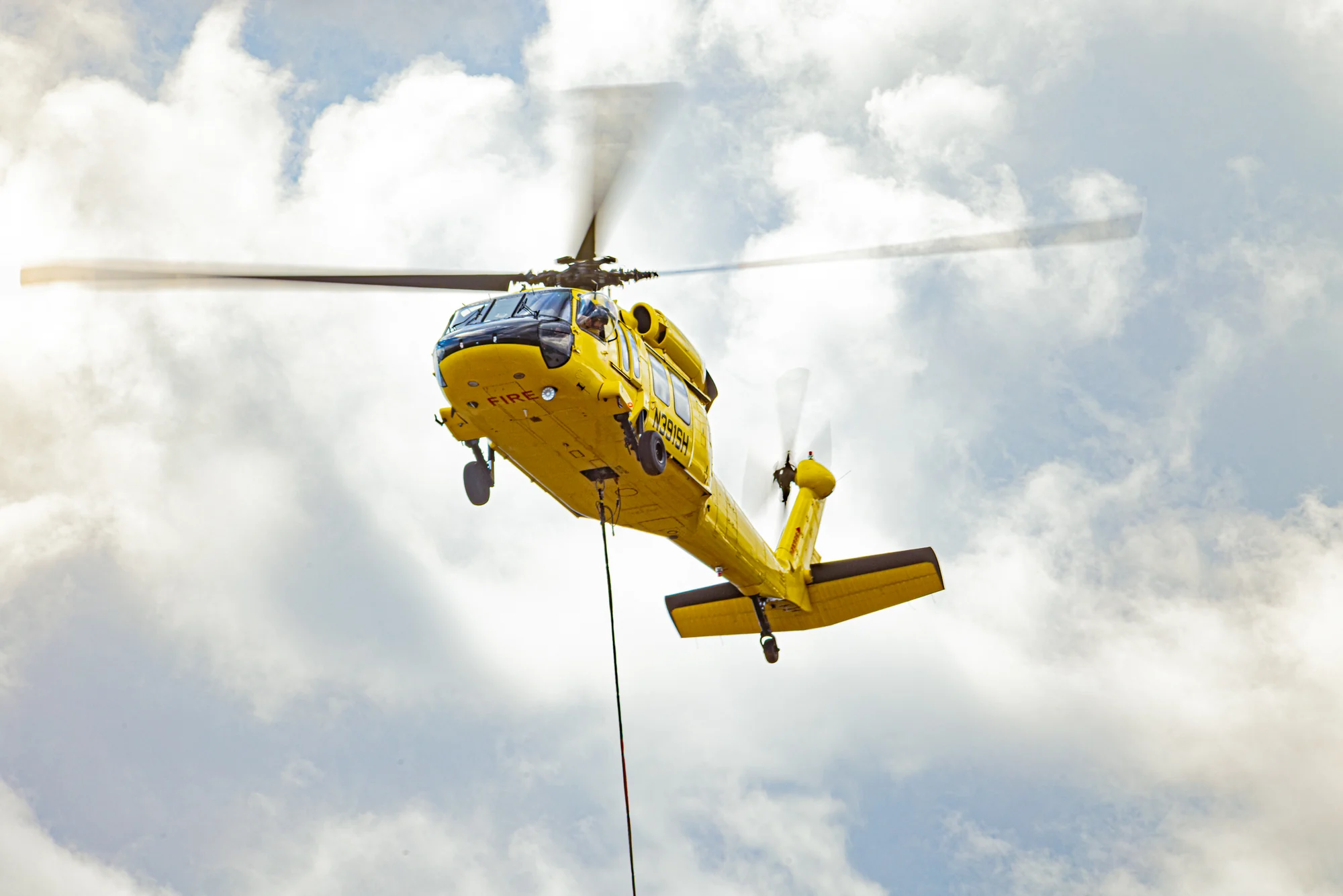 Yellow firefighting helicopter airborne with a longline extended beneath it against a cloudy sky.