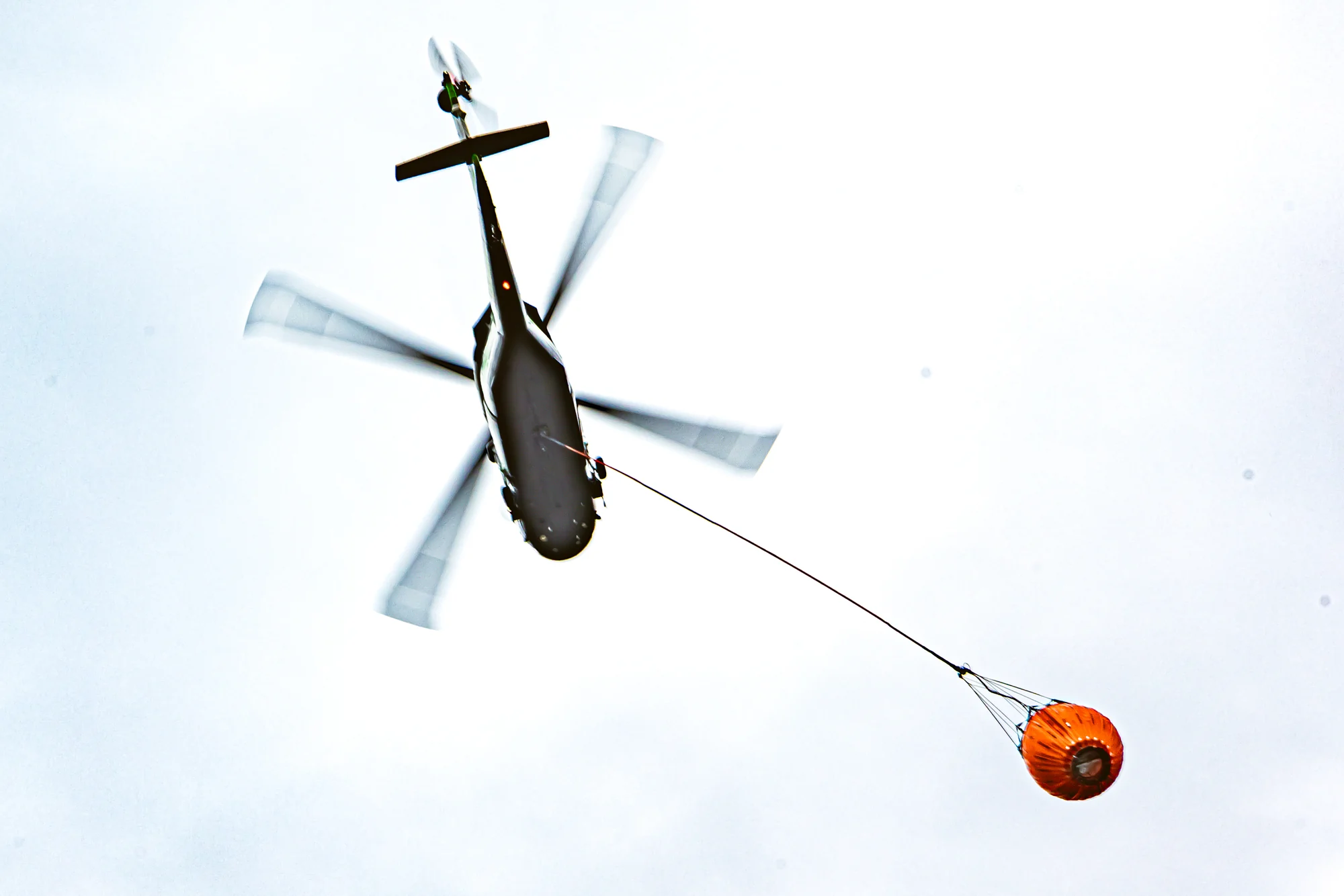 Firefighting helicopter viewed from below while carrying an orange water bucket on a longline.