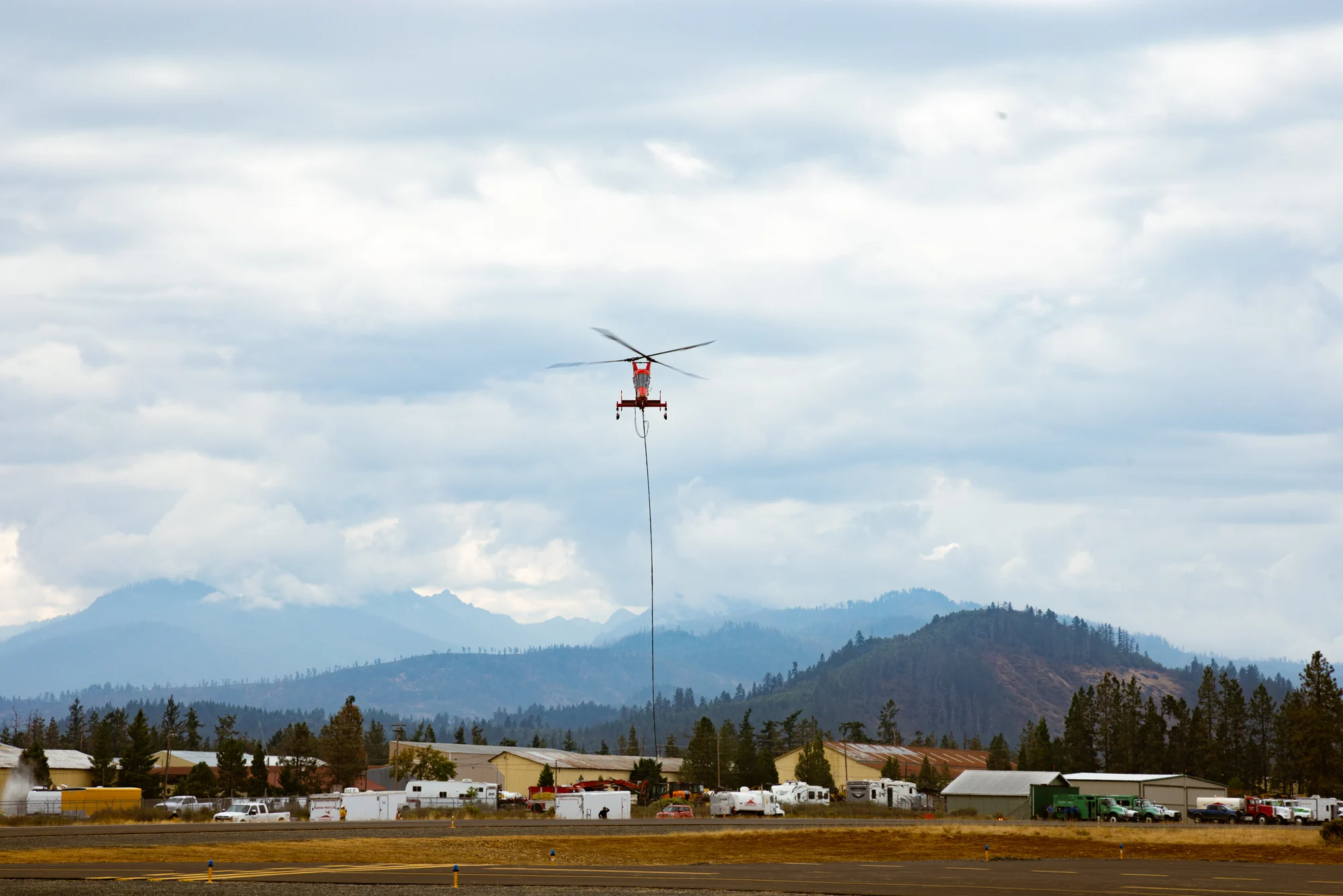 Firefighting helicopter carrying a longline high above an airport with forested mountains beyond.