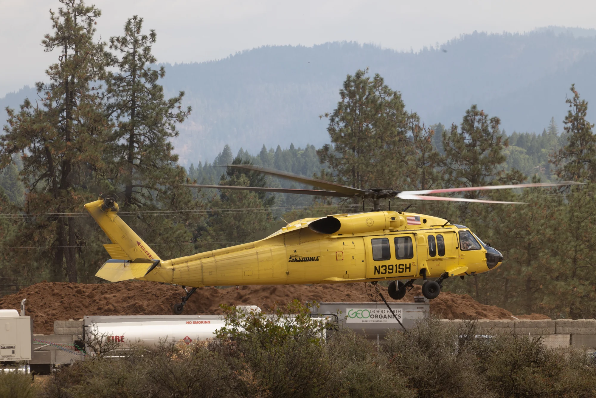 Yellow firefighting helicopter flying low near airport equipment with forested hills in the distance.