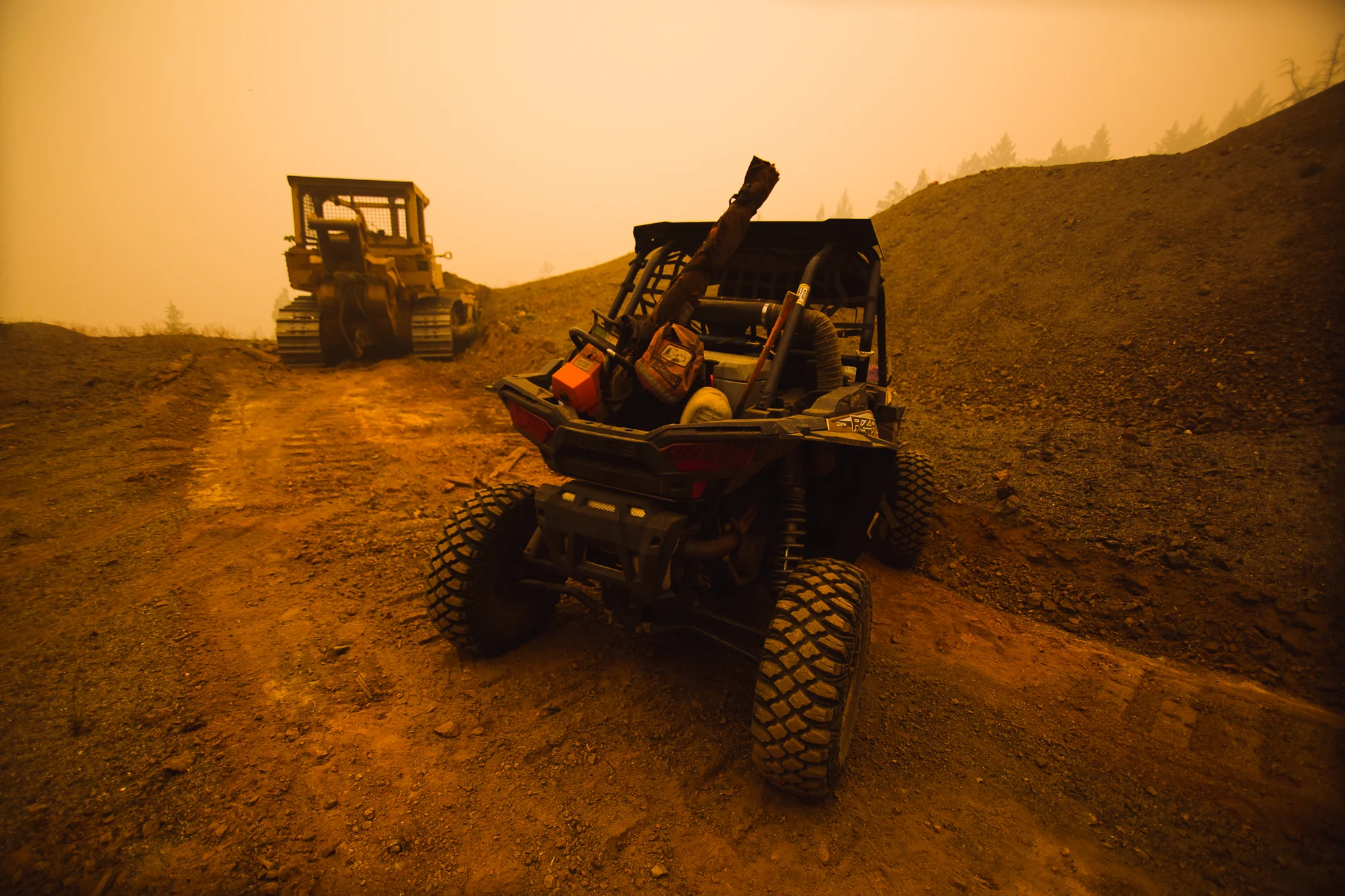 Off-road vehicle parked near a bulldozer beneath an orange, smoke-filled sky.